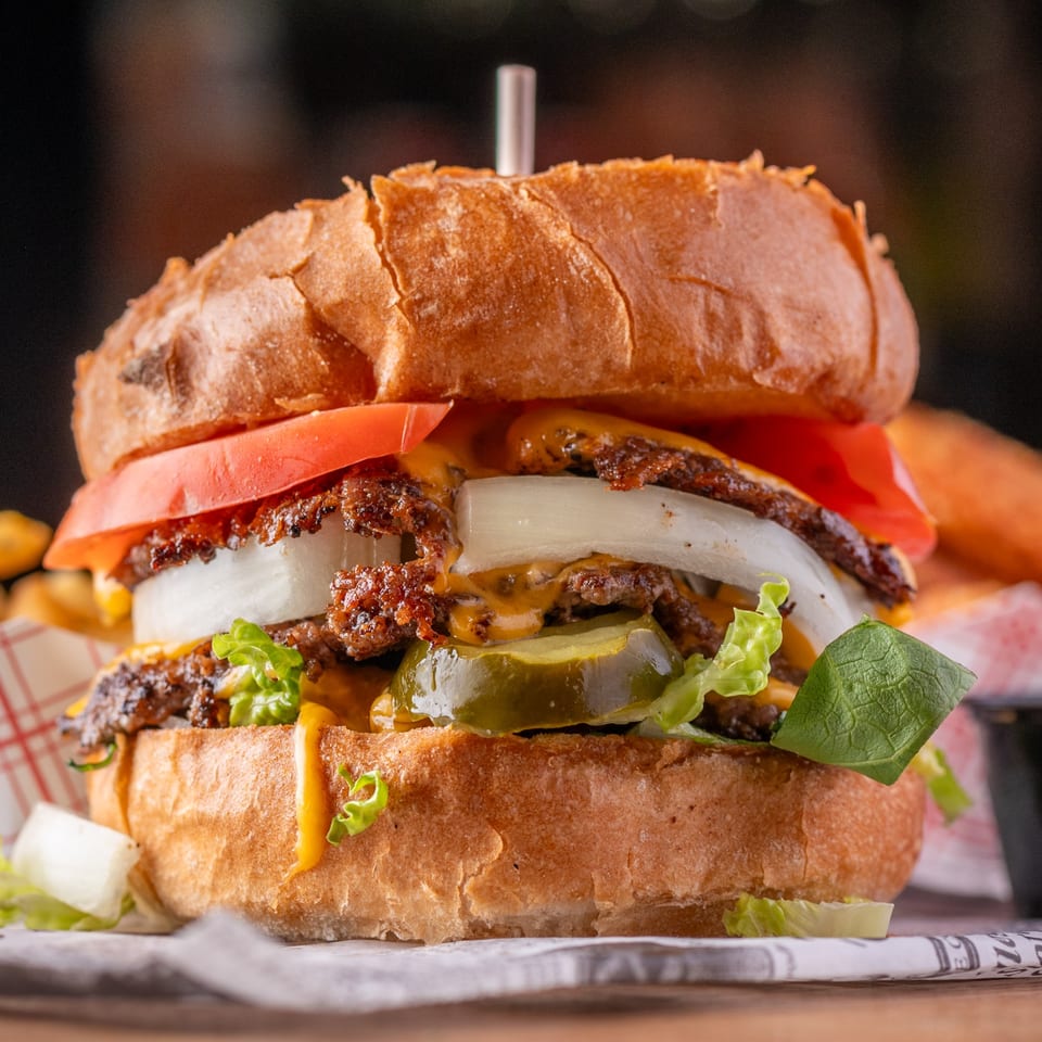 Close-up of a burger with two patties, melted cheese, onions, tomatoes, lettuce, and pickles.