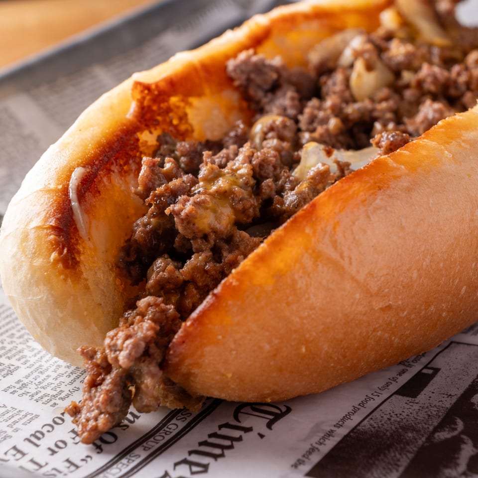 Close-up of a cheesesteak sandwich with ground meat and onions on a hoagie roll, served on newspaper.