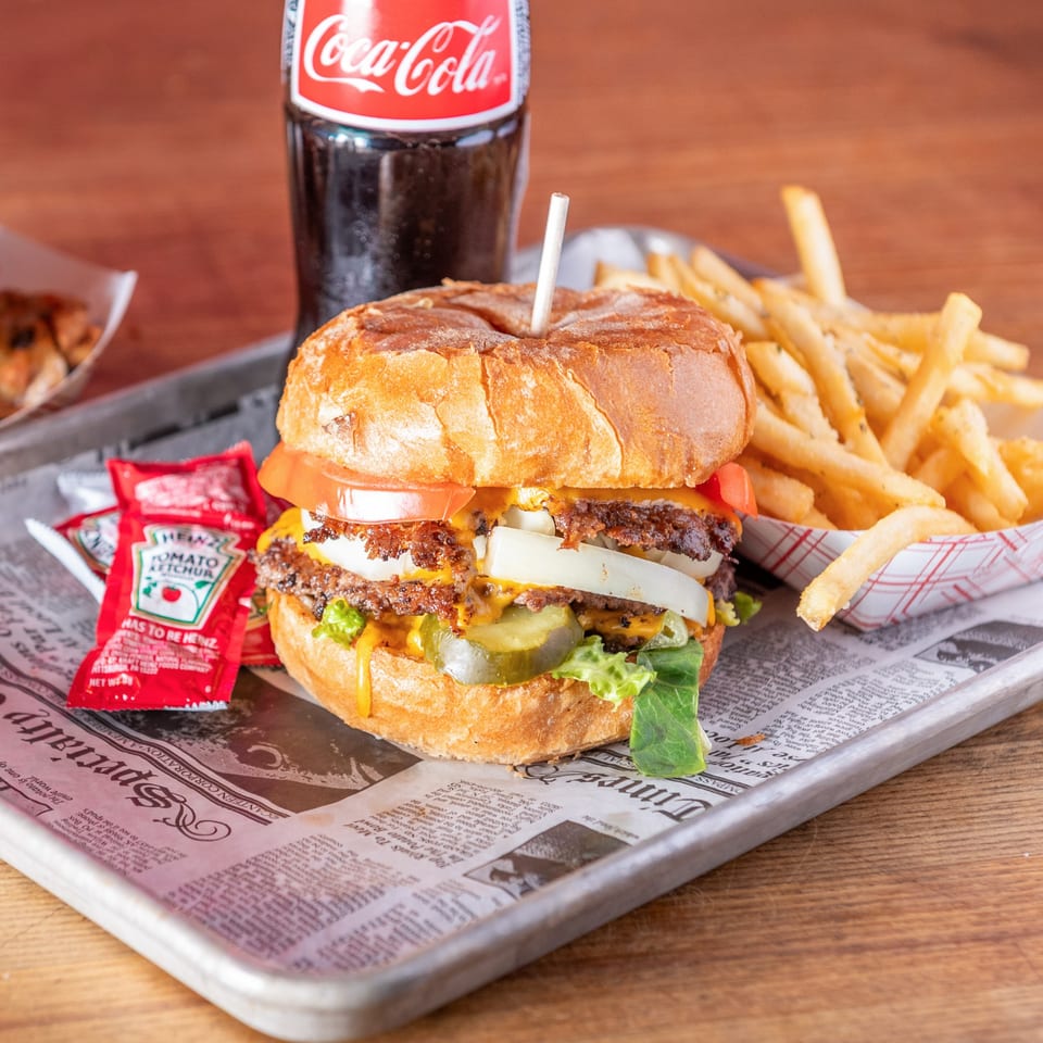 A double cheeseburger with fries, ketchup packets, and a Coca-Cola bottle on a newspaper-lined tray.