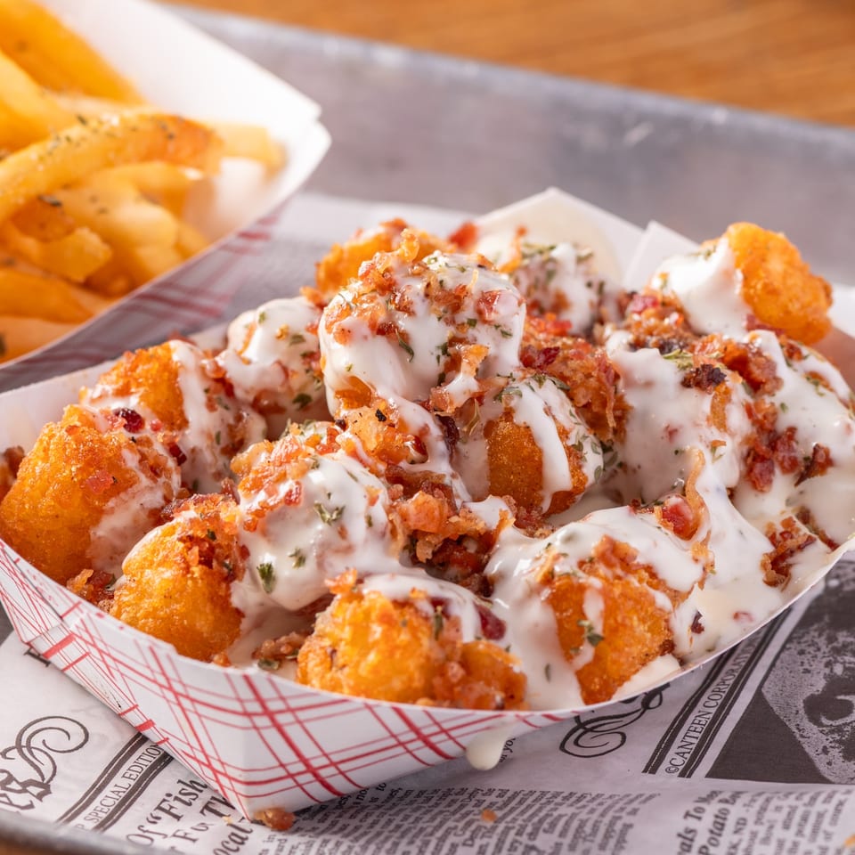 Golden fried cheese curds topped with white sauce, bacon bits, and herbs, next to fries.