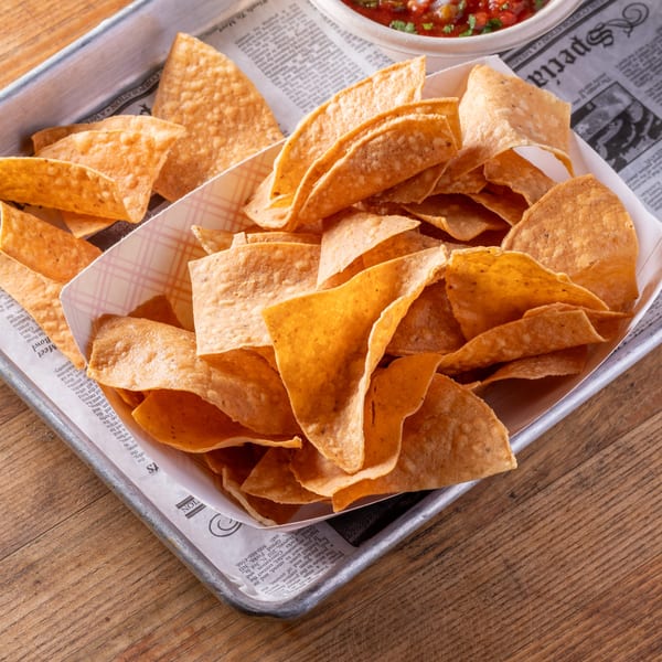 Crispy tortilla chips in a paper-lined metal tray, with a small bowl of salsa in the background.