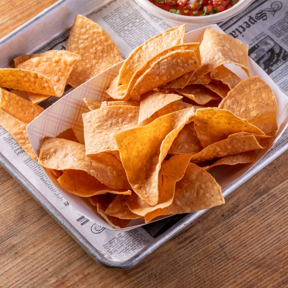 Crispy tortilla chips in a paper-lined metal tray, with a small bowl of salsa in the background.