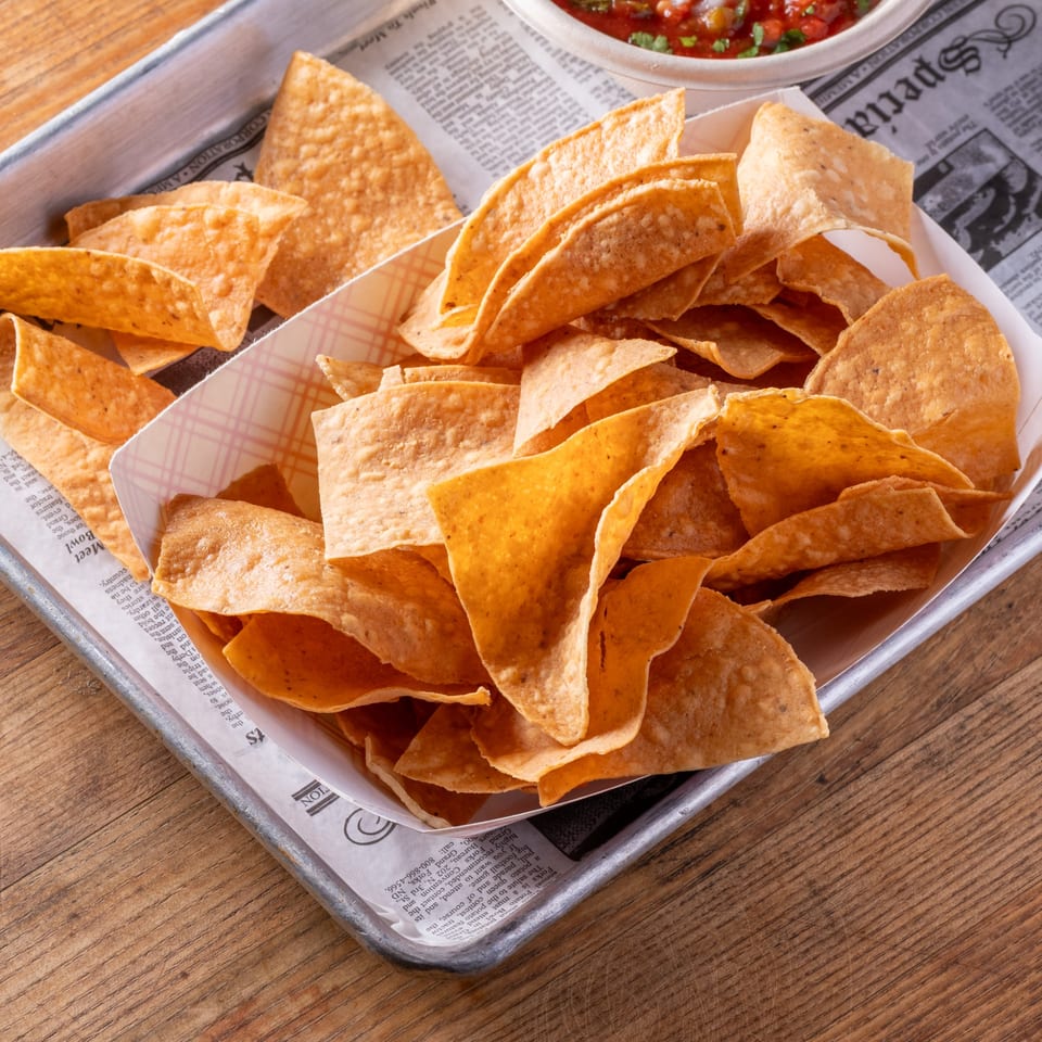 Crispy tortilla chips in a paper-lined basket on a metal tray, with salsa in the background.