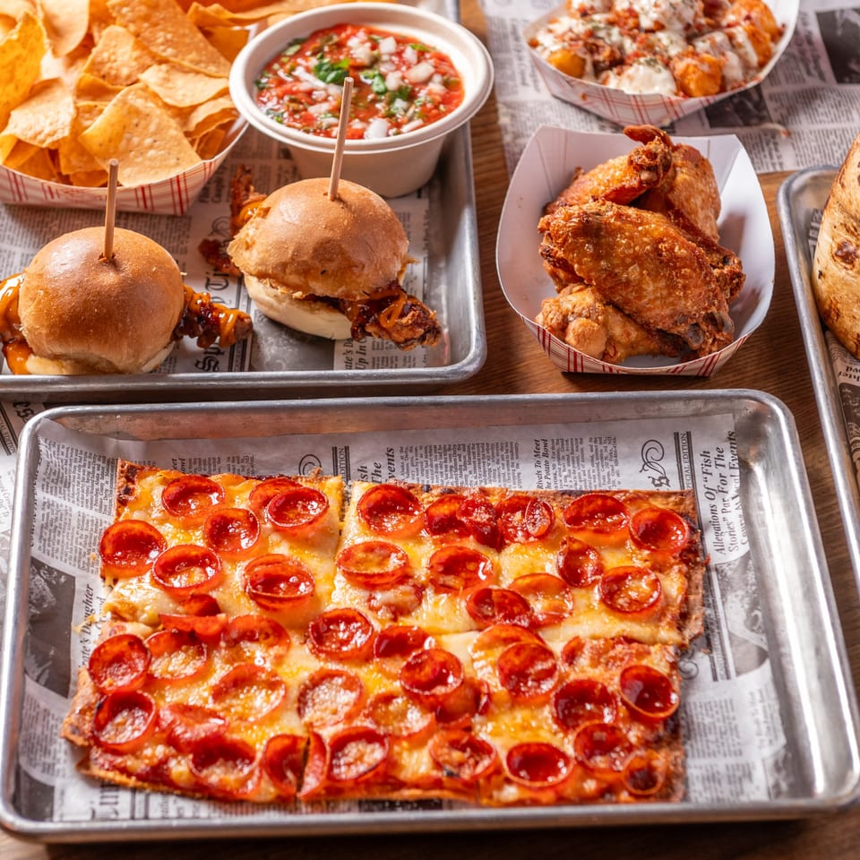 A pepperoni pizza, chicken wings, sliders, chips, and salsa are arranged on metal trays.